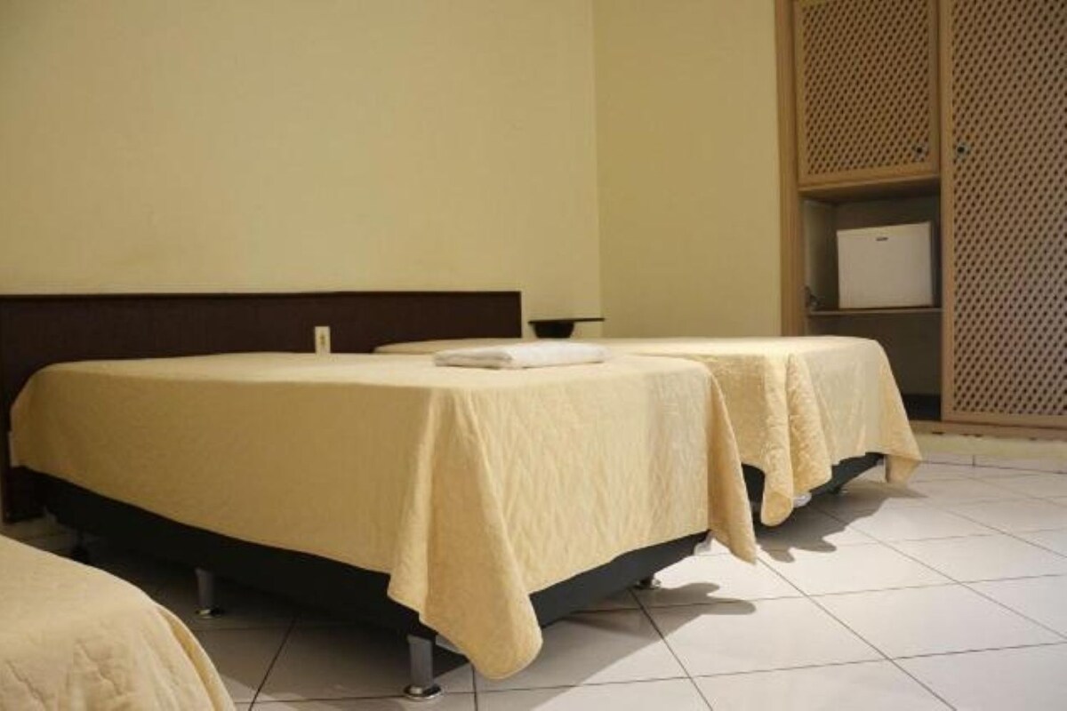Una habitación de hotel con dos camas, cada una cubierta con una manta color beige, y un mueble pequeño en la esquina.
