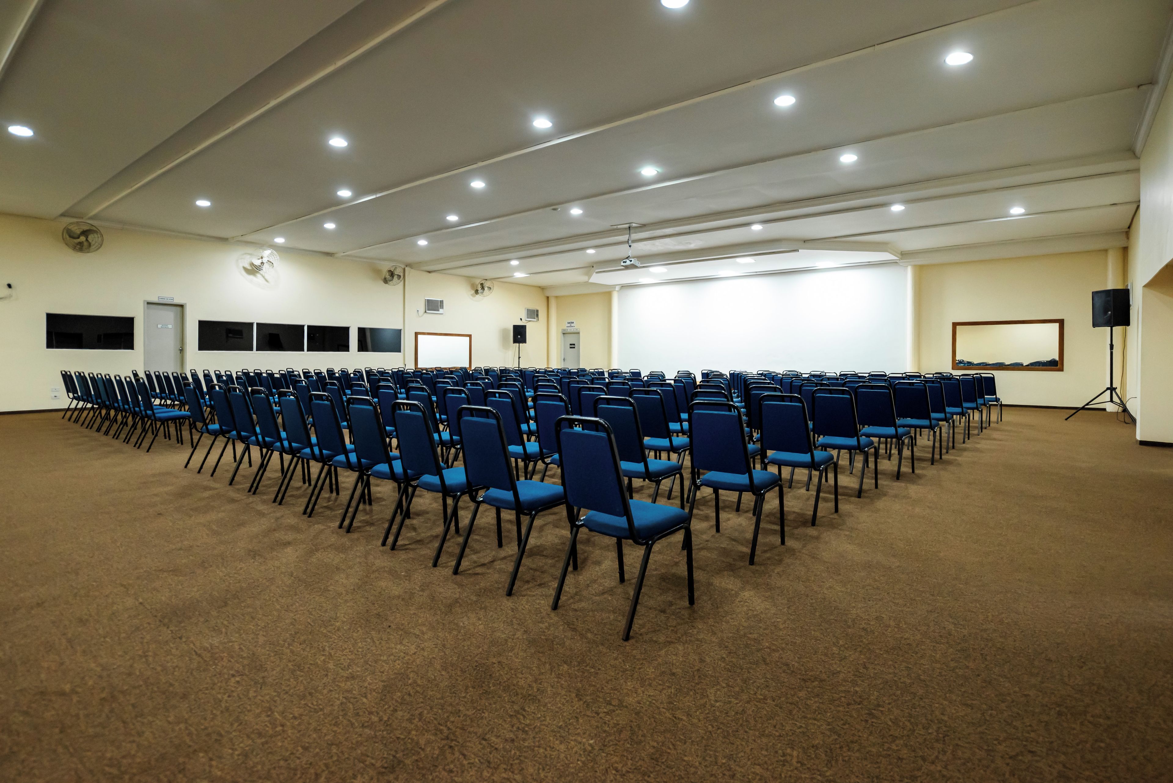 Uma sala de conferências com fileiras de cadeiras azuis, um quadro branco e uma tela de projetor.
