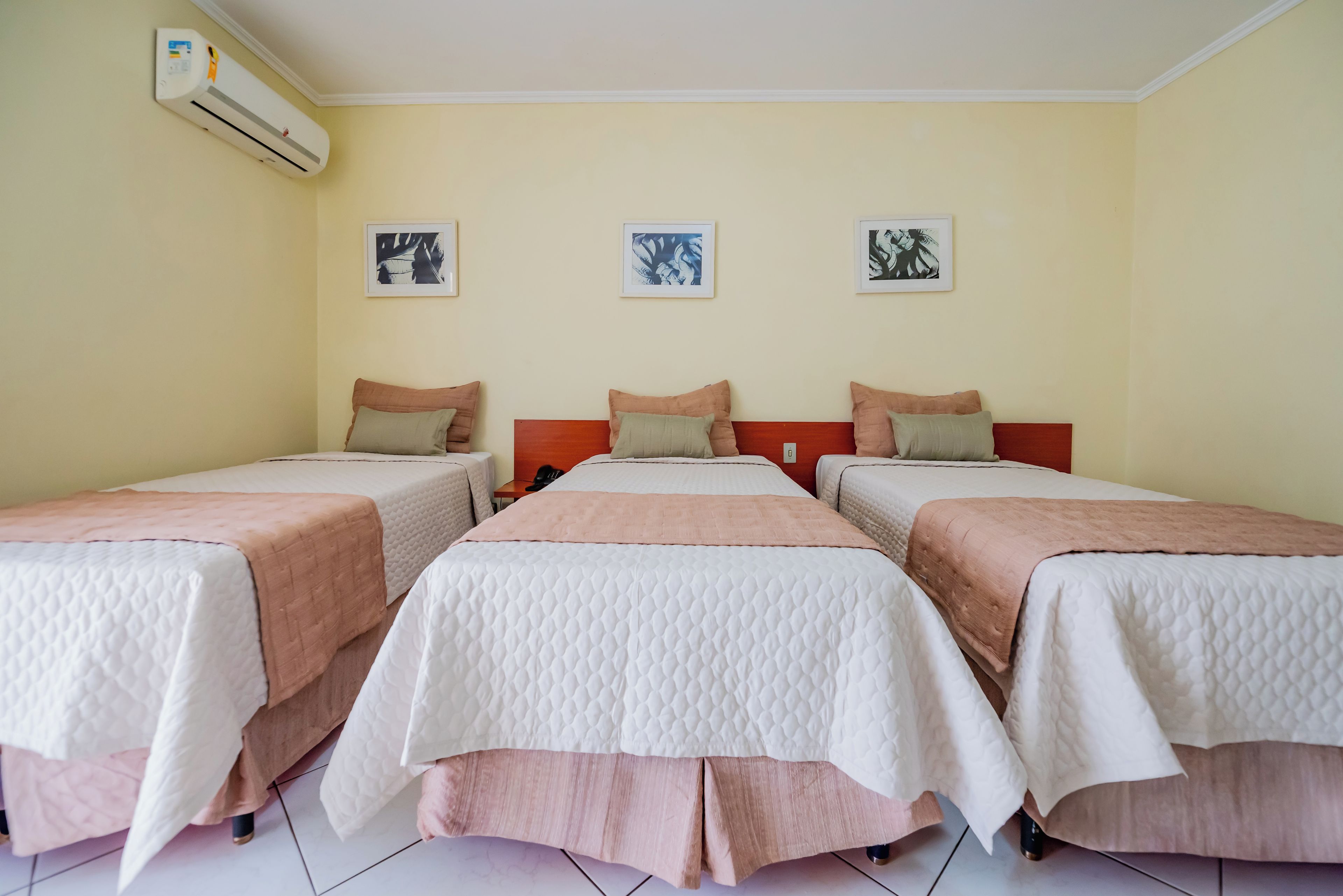 Quarto de hotel com três camas de solteiro, cada uma com colcha branca e rosa, e ar condicionado fixado na parede.
