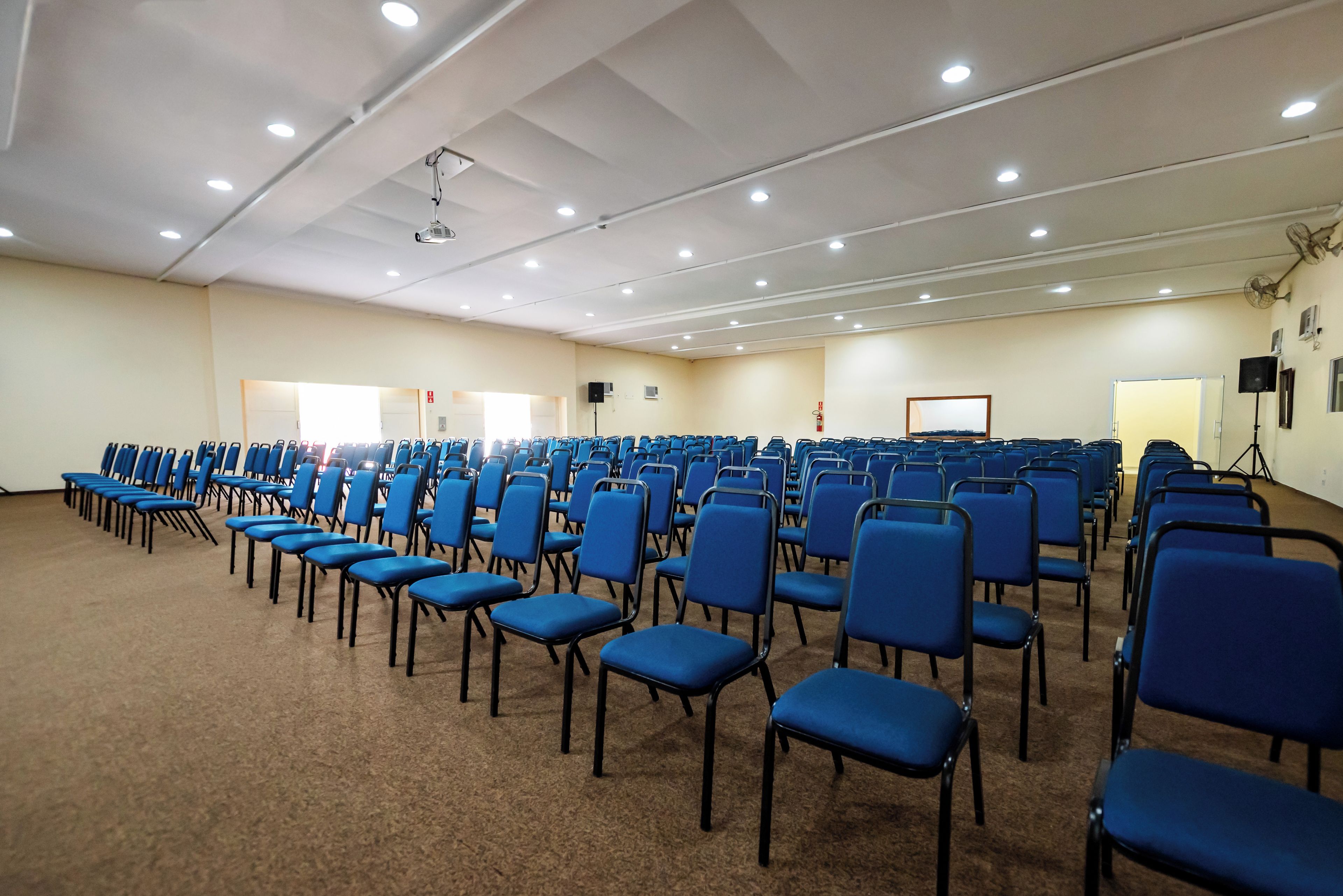 Un salón de conferencias con filas de sillas azules, un techo blanco con iluminación empotrada y una pantalla de proyector.