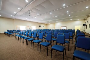 A conference room with rows of blue chairs, a white ceiling with recessed lighting, and a projector screen.