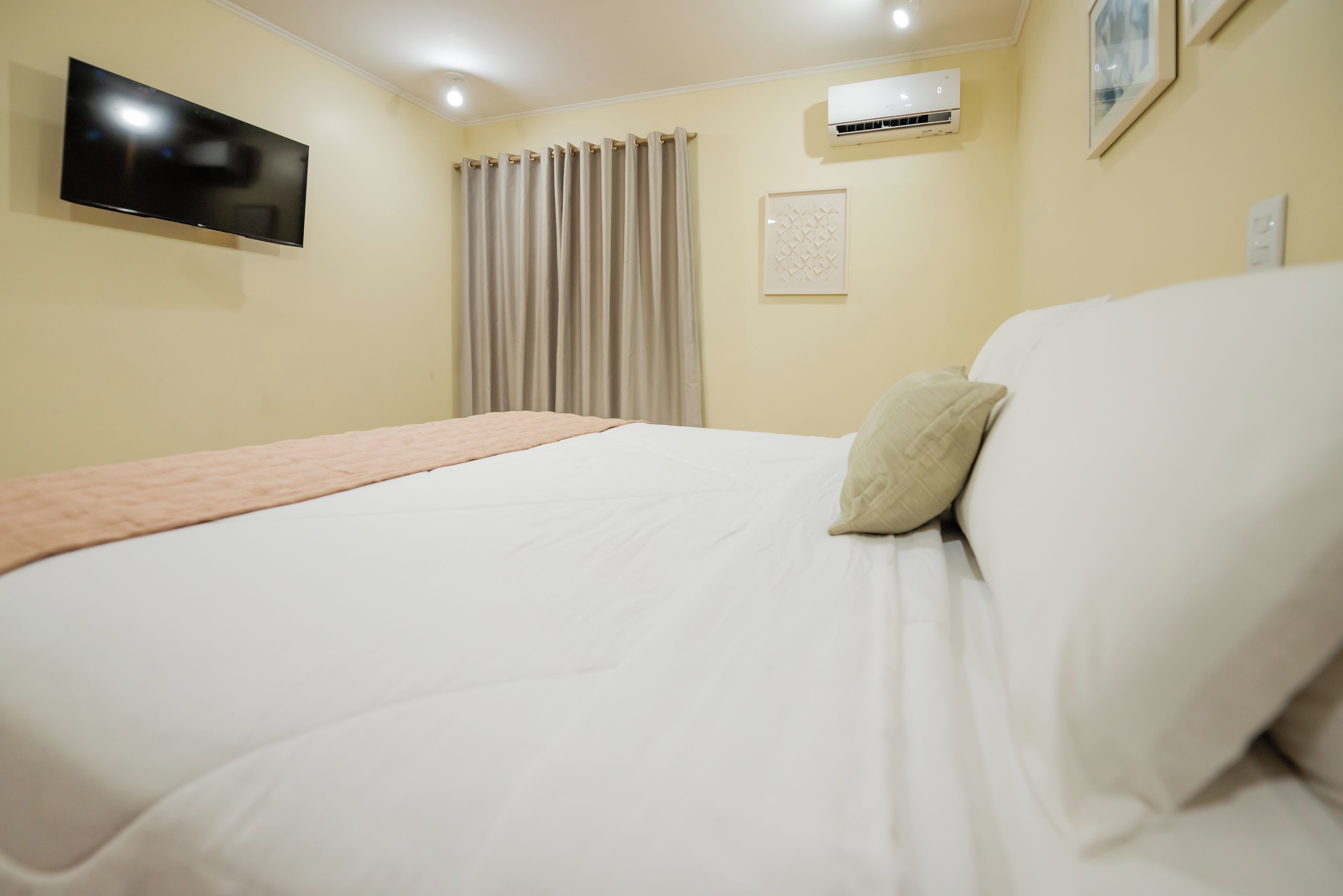 A hotel room with a bed, a flat-screen TV mounted on the wall, an air conditioning unit, and a window with curtains.