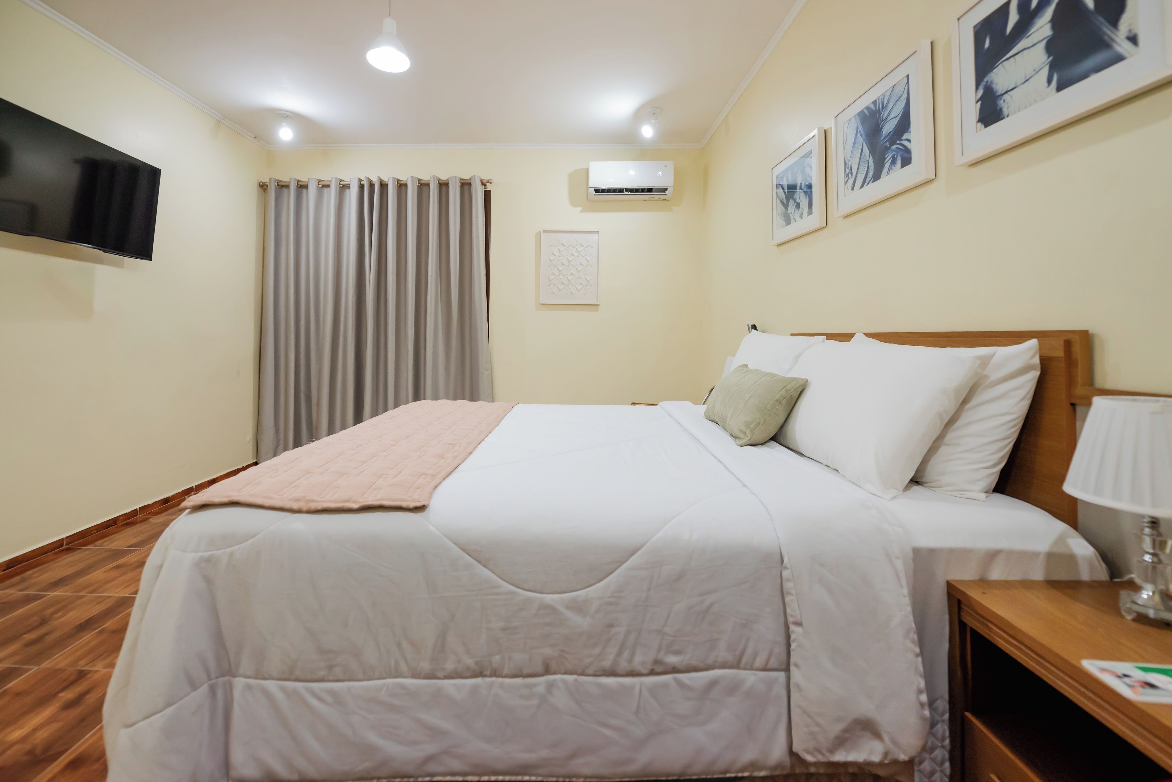A hotel room with a bed, a flat-screen TV mounted on the wall, a ceiling light, and a window with curtains.