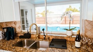 A kitchen with granite countertops, a double sink, a wine bottle, and a view of a backyard with a pool and potted plants.