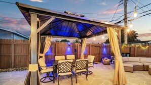 A covered outdoor patio with a table and chairs, a cushioned seating area, and decorative lighting.