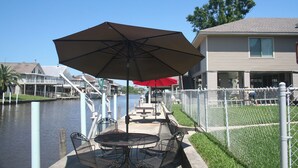 A patio with a table and chairs, a large umbrella, and a view of a water body and buildings in the background. - Camp Gator #2 (Barataria)