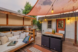 Un espacio al aire libre acogedor con terraza de madera, banca acolchada, mobiliario de mimbre y una zona cubierta con guirnaldas de luces.