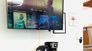 Un soggiorno con una TV a schermo piatto montata a parete che mostra un film. Sotto la TV, un mobiletto in legno con una macchina da caffè e una piccola pianta sopra.