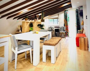 A dining area with a white table and benches, a kitchen in the background, and a wooden ceiling.