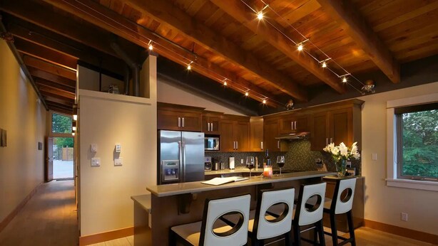 A modern kitchen with wooden beams, a central island with bar stools, stainless steel appliances, and a view of the outdoors. - Mariner's Loft Salt Spring Island - 2BDRM - Unit 201 (Salt Spring Island)