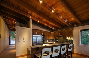 A modern kitchen with wooden beams, a central island with bar stools, stainless steel appliances, and a view of the outdoors. - Mariner's Loft Salt Spring Island - 2BDRM - Unit 201 (Salt Spring Island)