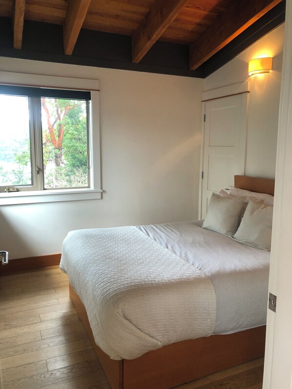 A bedroom with a wooden bed, a window with a view of trees, and a wall-mounted light fixture. - Mariner's Loft Salt Spring Island - 2BDRM - Unit 201 (Salt Spring Island)