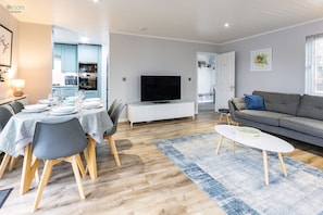 A modern living room with a dining area, a grey sofa, a flat-screen TV, and a kitchen in the background. - Windrush Lake 41, Kingfisher House - Lakeside Lodge, On-site Activities, Hot Tub (South Cerney)