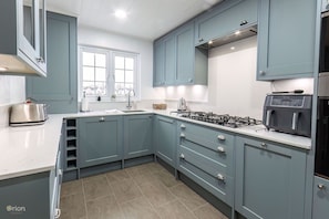 A kitchen with teal cabinets, a gas stove, a microwave, and a window with a view of houses. - Windrush Lake 41, Kingfisher House - Lakeside Lodge, On-site Activities, Hot Tub (South Cerney)