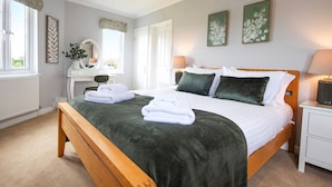 Une chambre bien rangée, avec un lit en bois, du linge de lit blanc et un plaid vert. On y trouve une table de chevet avec une lampe, un miroir et une plante. La pièce est dotée d’une fenêtre avec des rideaux et d’œuvres d’art accrochées au mur.