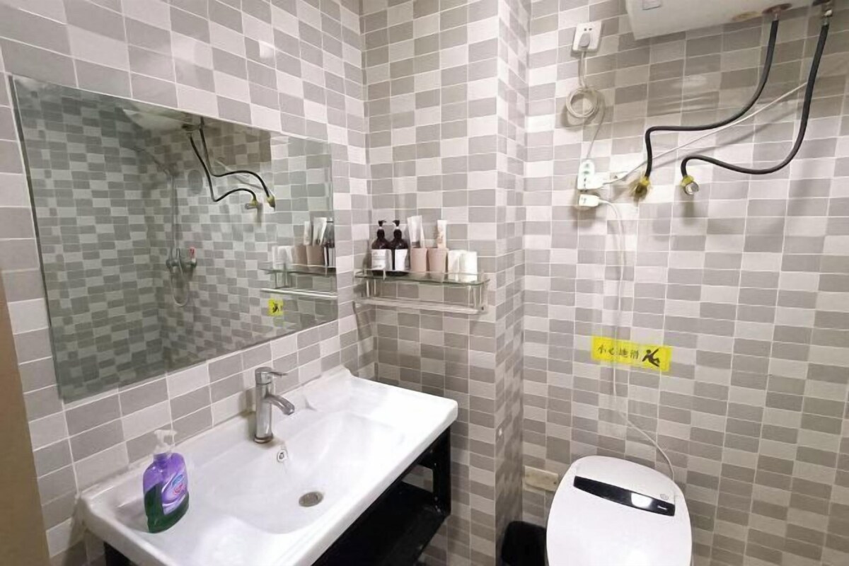 Ein Badezimmer mit einem weißen Waschbecken, einem Spiegel und einer Glasdusche. Auf einem Regal stehen Pflegeprodukte und ein Schild ist an der Wand angebracht.
