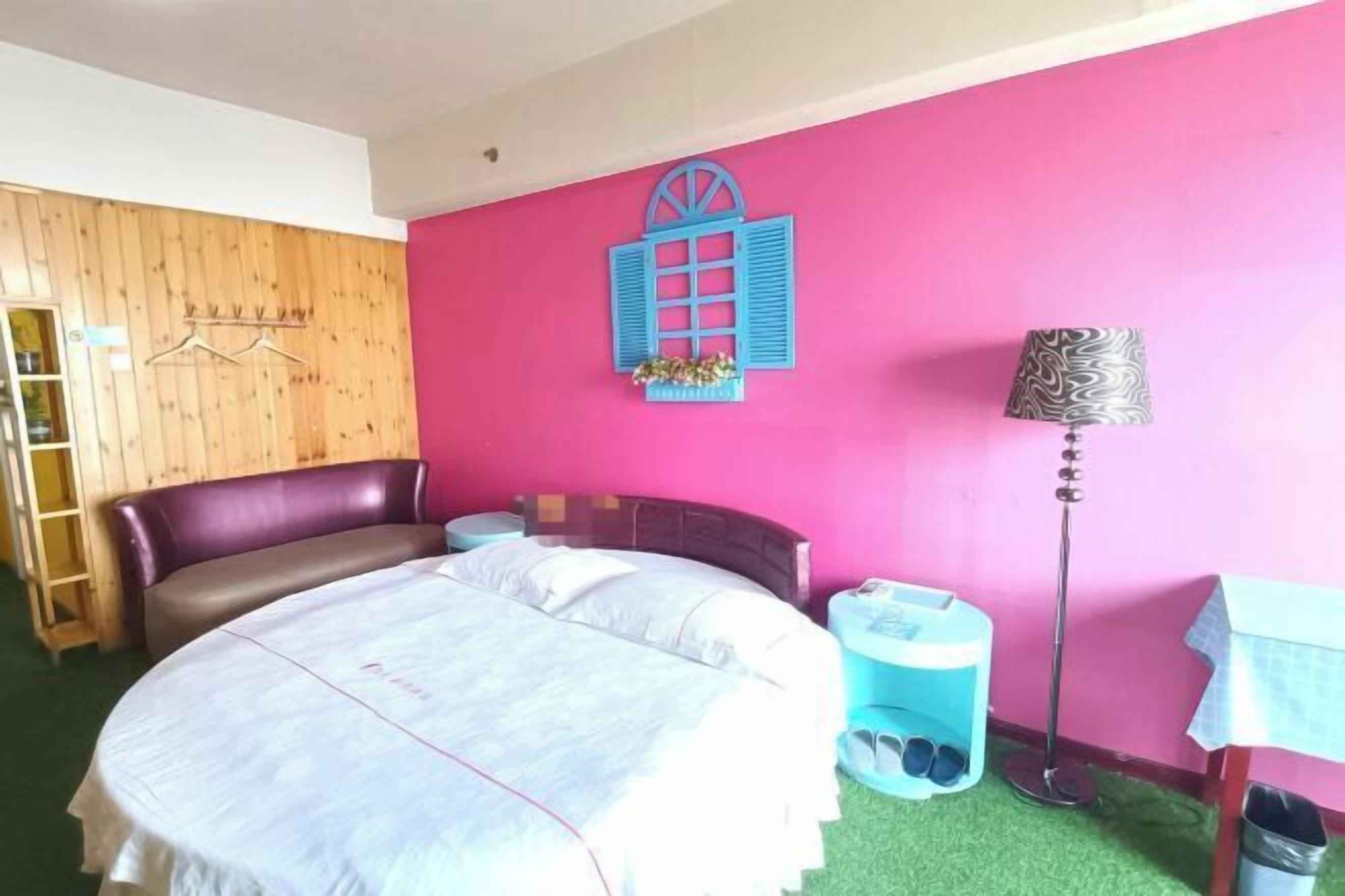 A bedroom with a bed, a wooden wardrobe, a pink wall, a blue window, a lamp, and a trash bin.