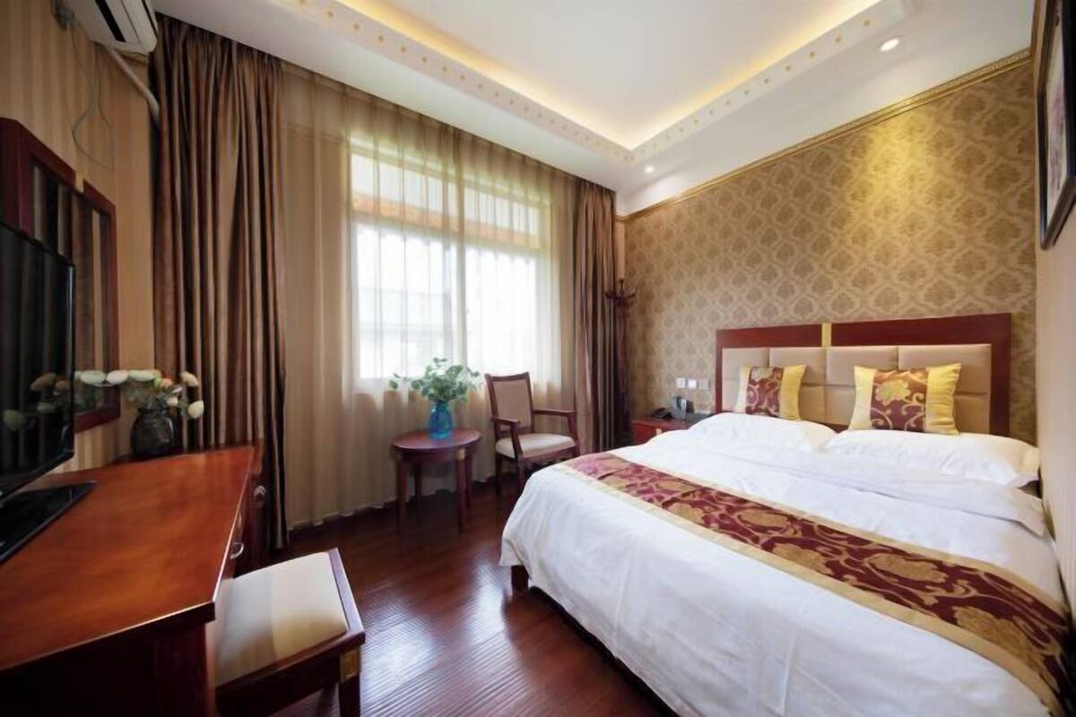 A hotel room with a large bed, a desk, and a chair. There is a window with curtains, a small table with a vase, and a wall-mounted TV.