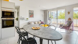 A modern kitchen with a built-in oven and microwave, a dining table with chairs, a sofa with cushions, and a balcony with a chair.