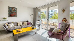 A modern living room with a grey sofa, a yellow coffee table, and a purple armchair. There is a large window with a view of buildings and trees.