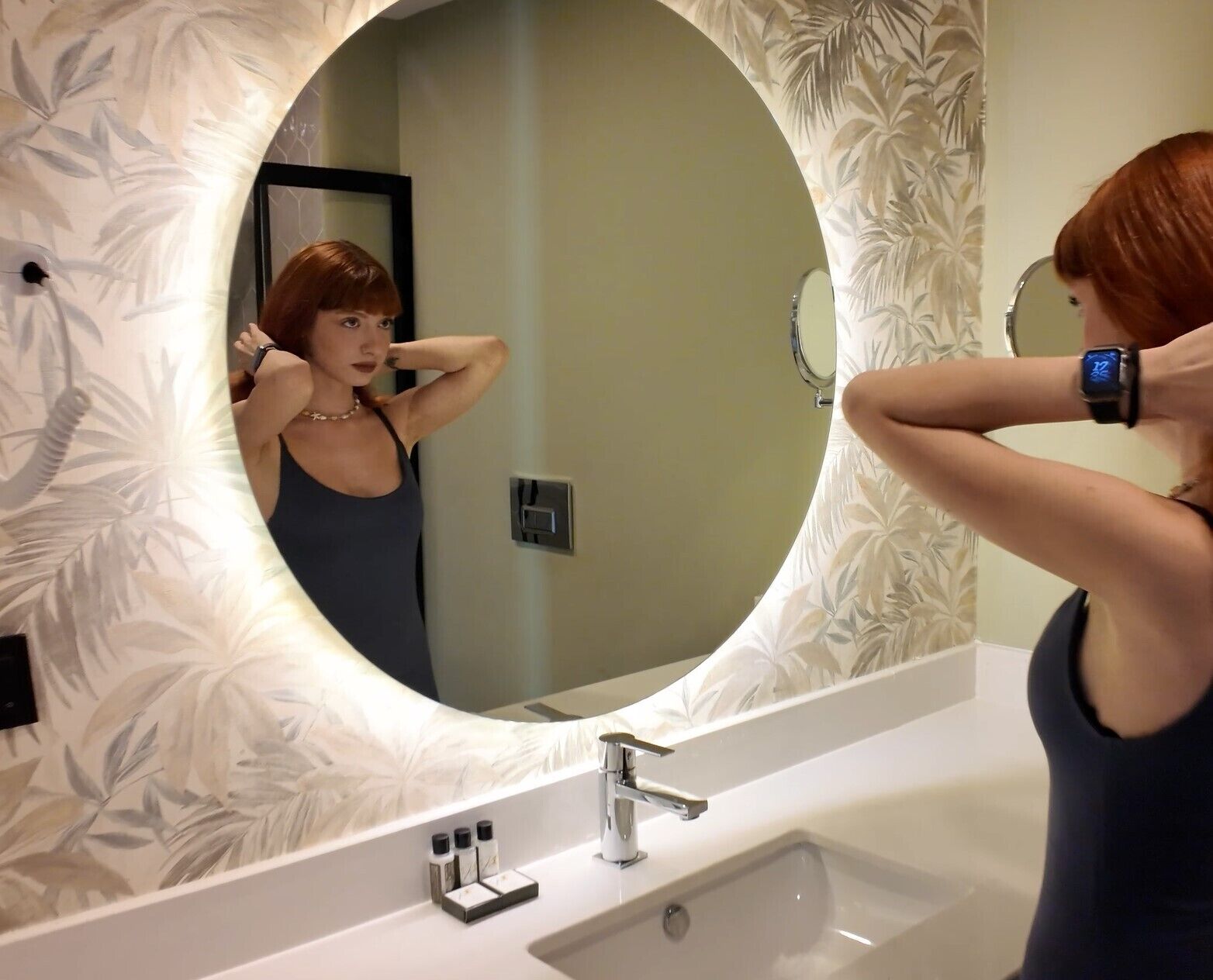 A bathroom with a large mirror, a white sink, and a floral-patterned wall.