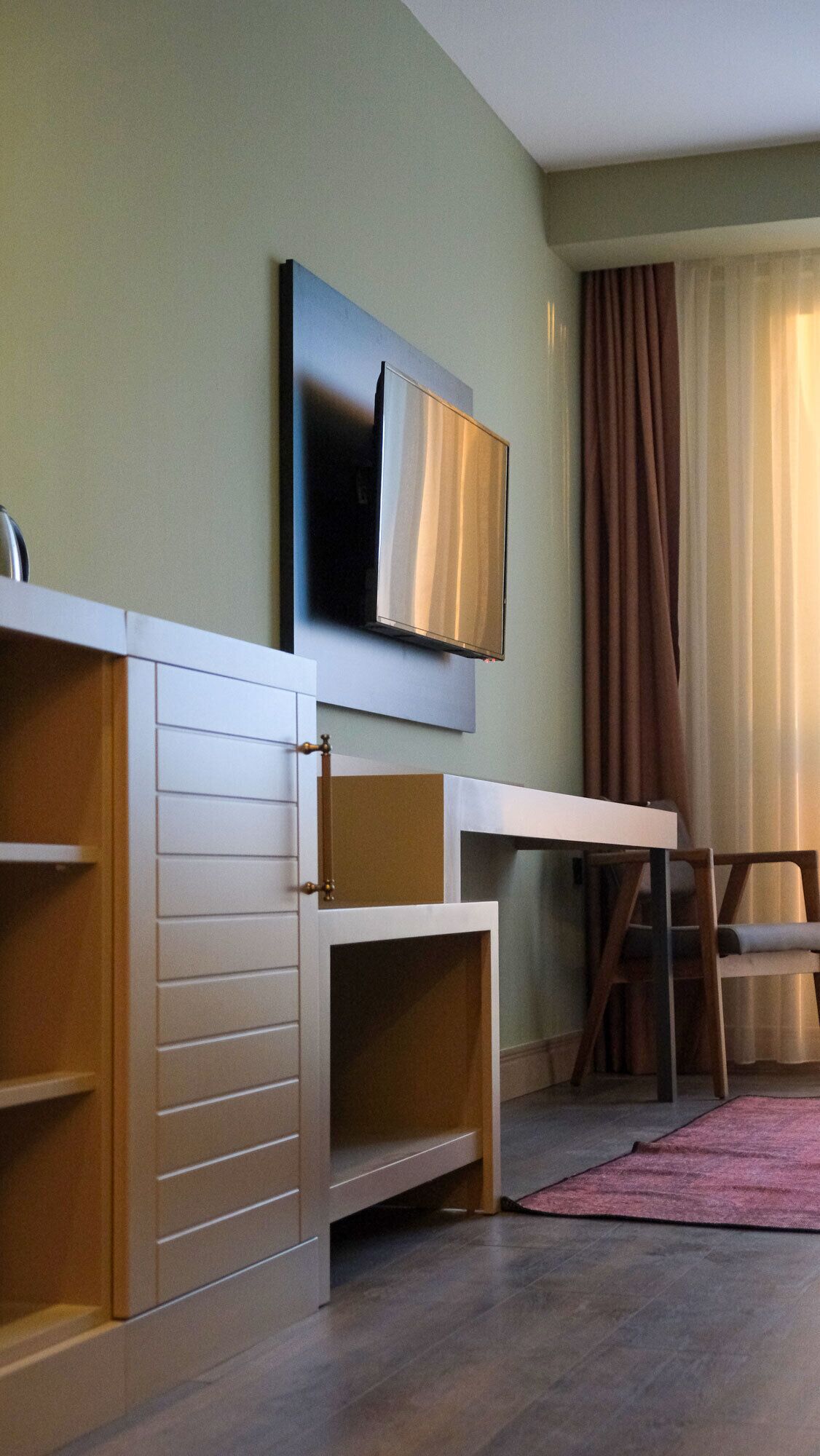 A modern living room with a flat-screen TV mounted on the wall, a wooden cabinet with drawers, a dining table, and a chair.