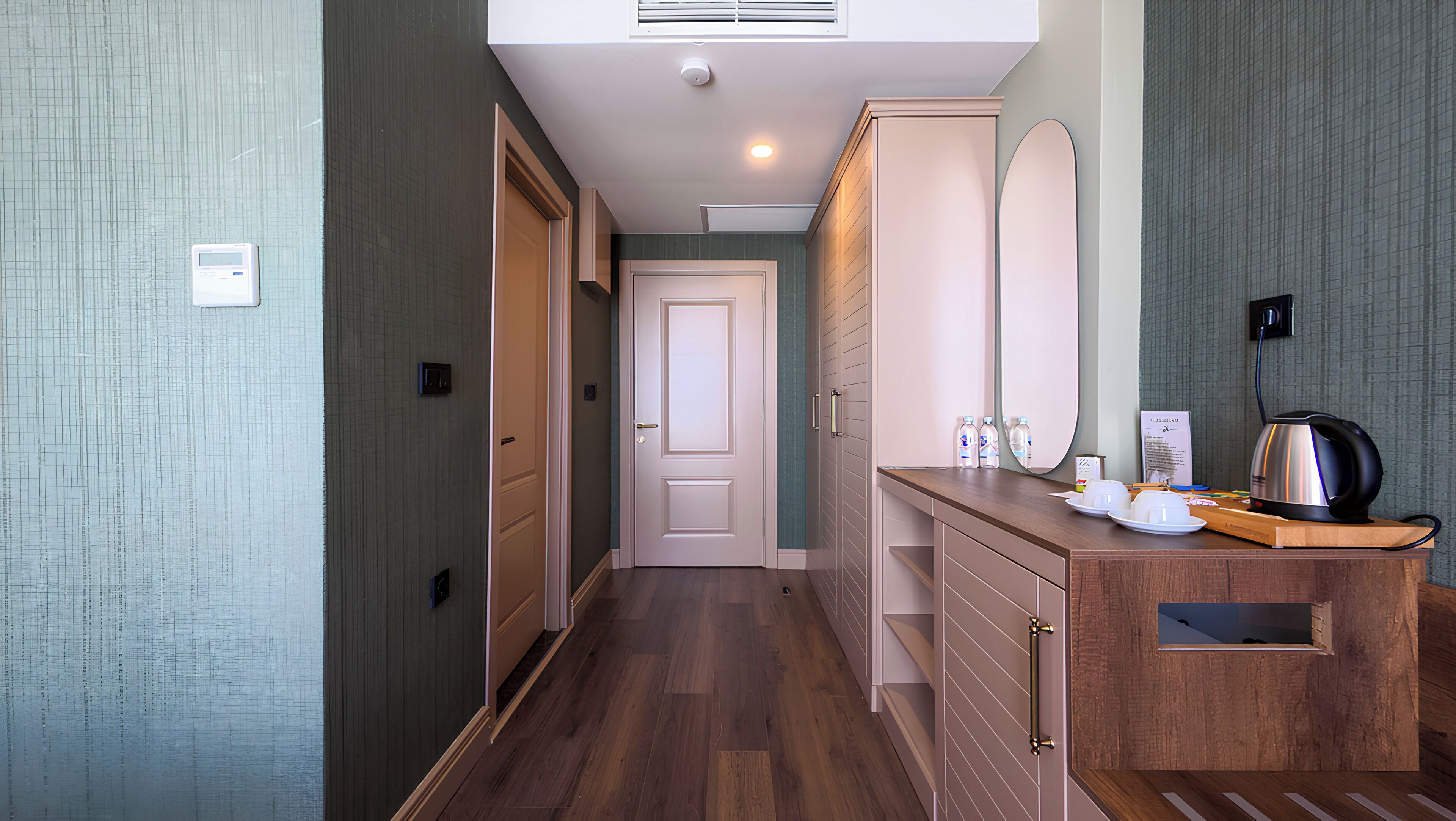 A hallway with wooden flooring, a kitchenette with a kettle, and a door leading to another room.