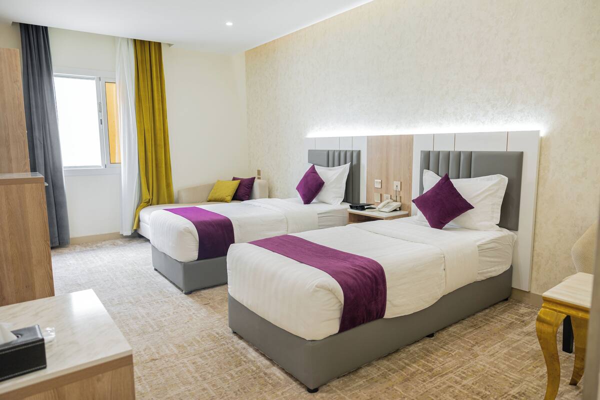 A hotel room with two beds, a bedside table, a nightstand, a lamp, a chair, and a window with curtains.