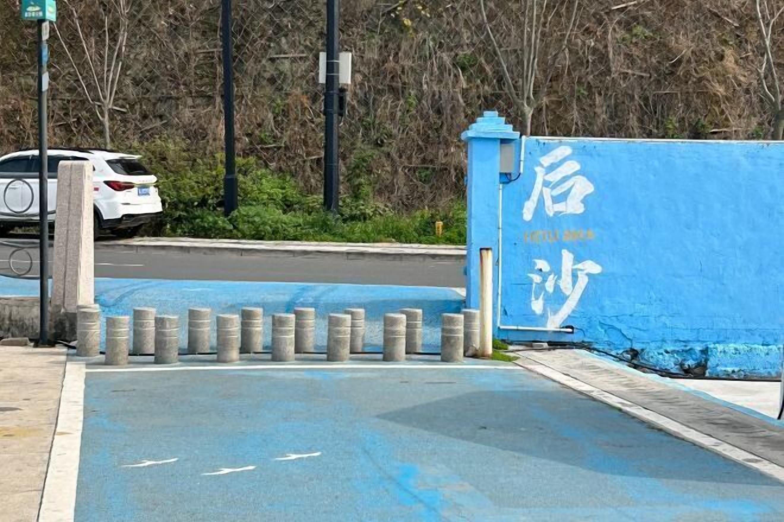 Una verja azul con caracteres chinos blancos, un coche blanco estacionado en la calle y separadores a lo largo de la acera.