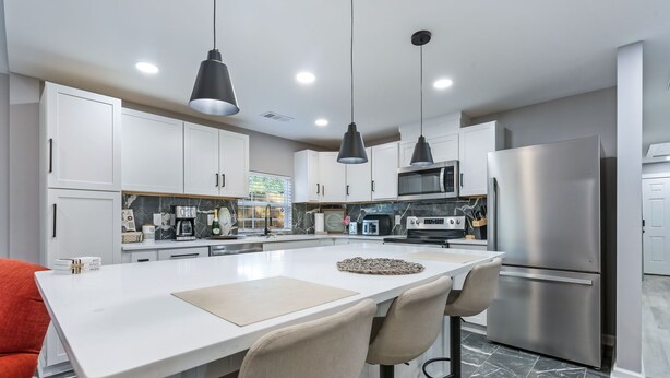 Una cucina moderna con mobili bianchi, un'isola centrale con sgabelli da bar, elettrodomestici in acciaio inossidabile e illuminazione a sospensione.