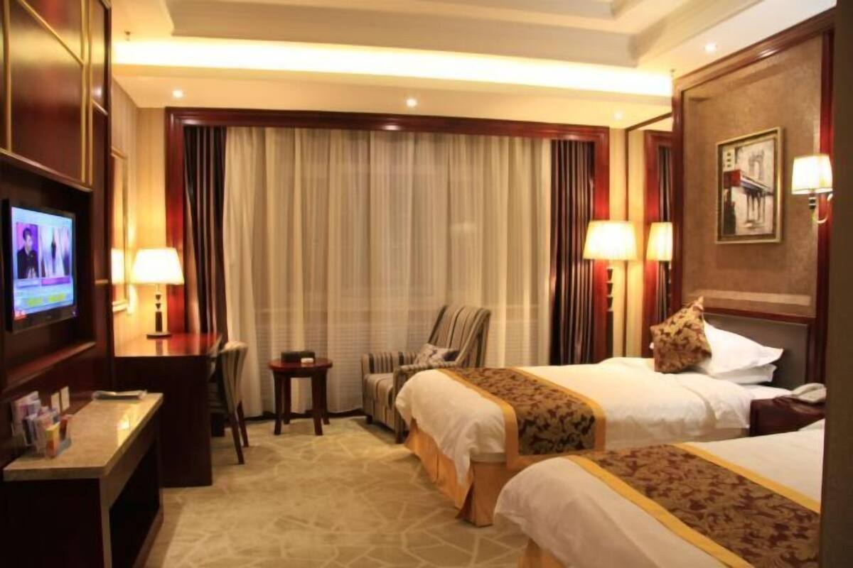 A hotel room with two beds, a desk with a TV, a chair, a small table, and a wall-mounted lamp.