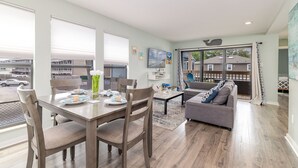 A modern living room with a dining area, a gray sofa, and a large window with a view of the street. - Private & Comfy Oasis: 5-star, Ocean View, Pool, Near Golf ~ Free Parking! (Myrtle Beach)
