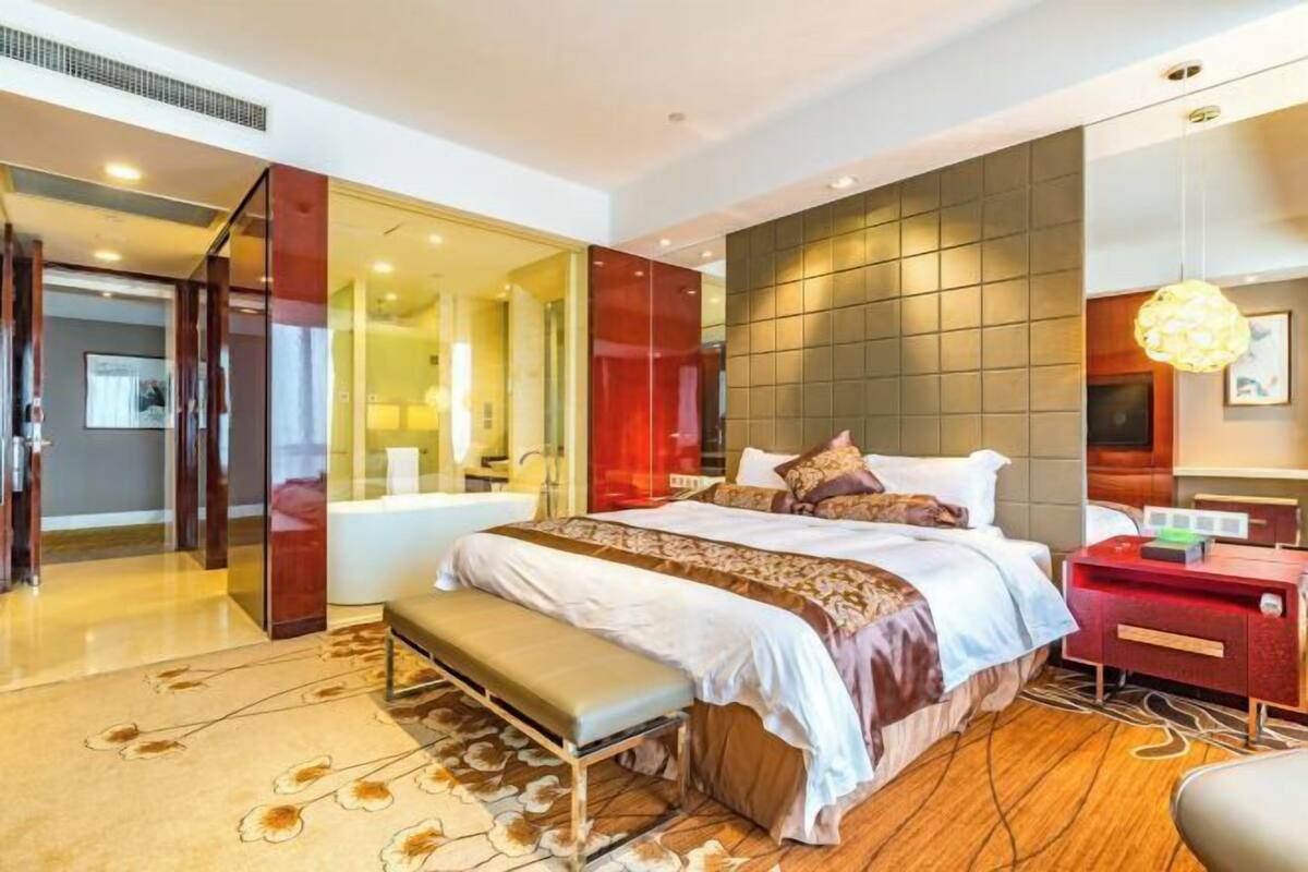 Ein modernes Hotelzimmer mit einem großen Bett, einer Sitzbank, einem Schreibtisch und einem durch eine Glastrennwand abgetrennten Badezimmer.
