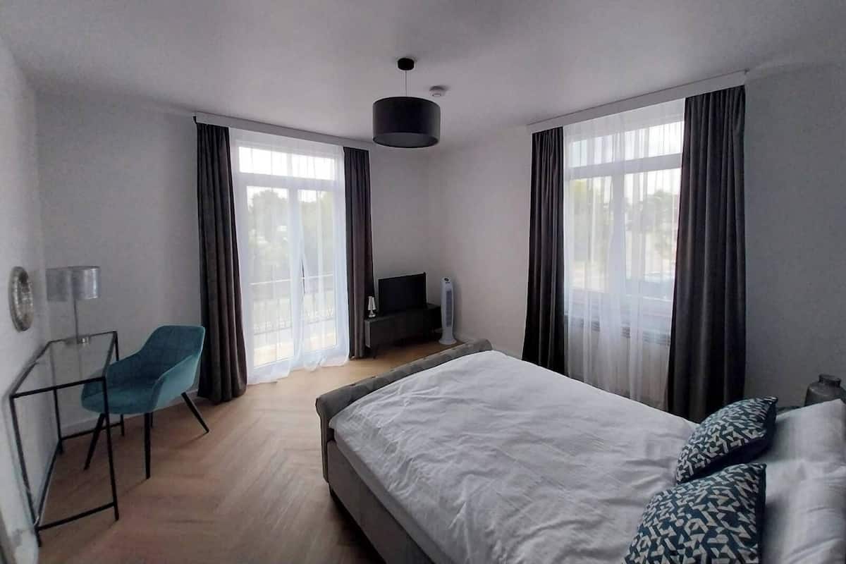 Una habitación de hotel moderna con una cama grande, un escritorio con silla, un televisor y ventanas amplias con cortinas.
