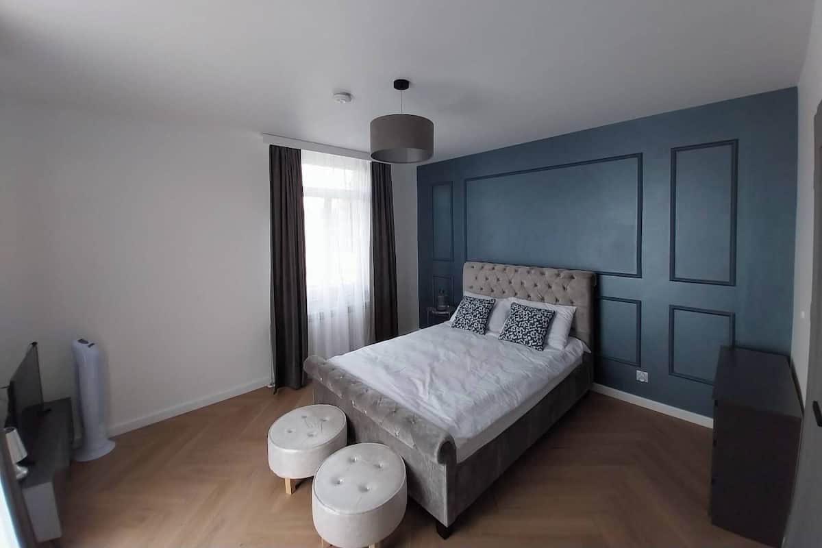 Un dormitorio moderno con una cama, dos otomanas, un aparador y un ventanal con cortinas.