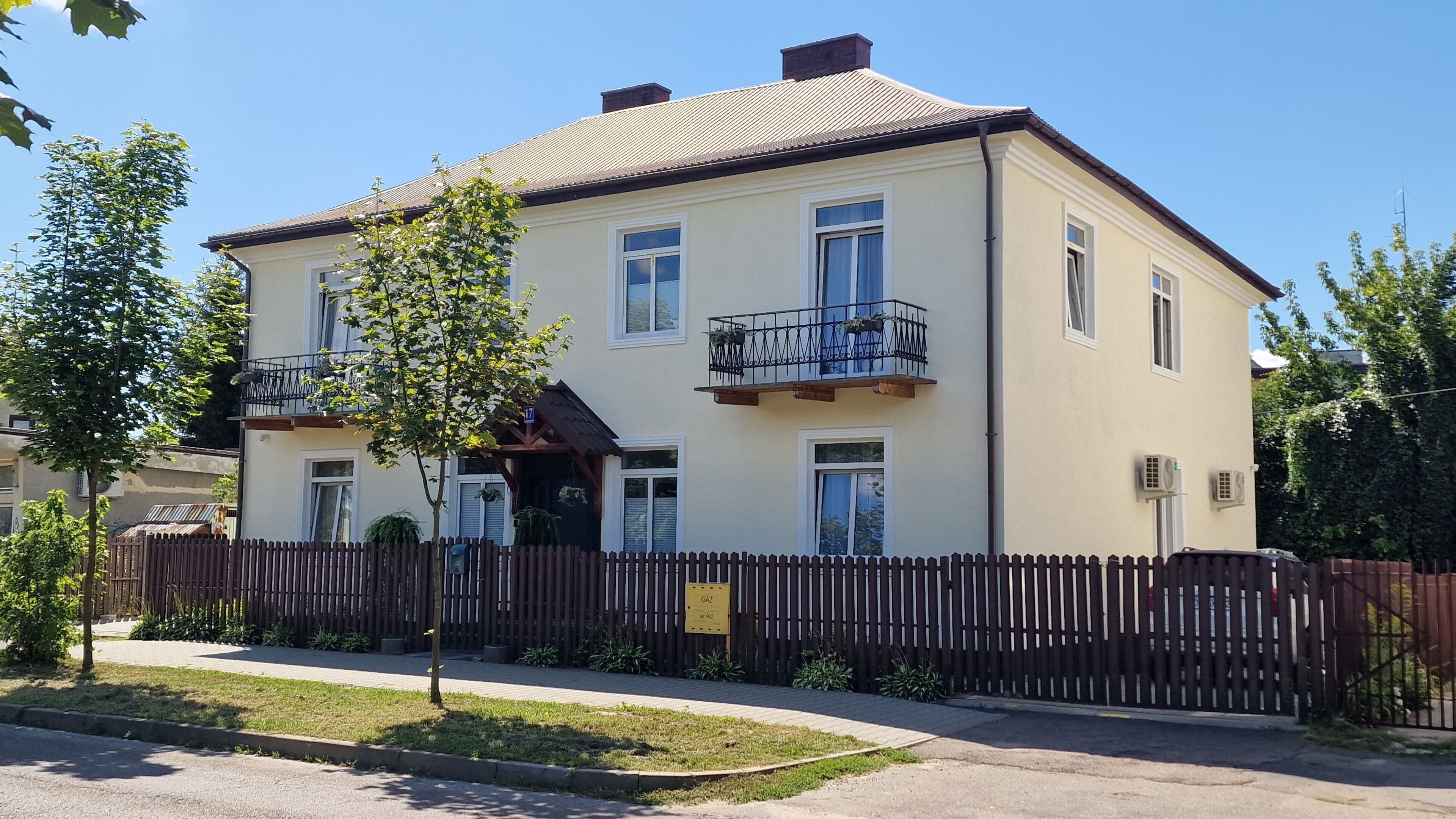 Ein zweigeschossiges Gebäude mit einem Balkon, einem Holzzaun und einem kleinen Baum im Vorgarten.