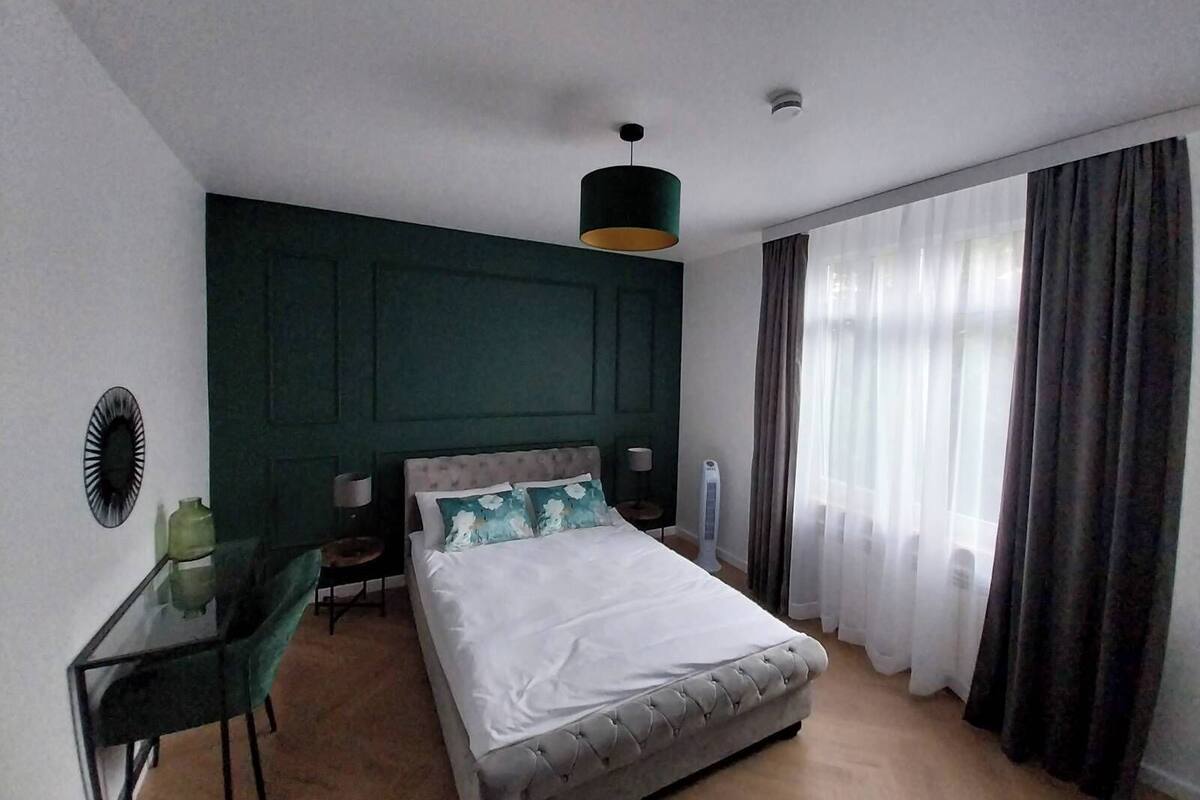Un dormitorio con cama, escritorio, silla, lámpara, calefactor y ventana con cortinas.
