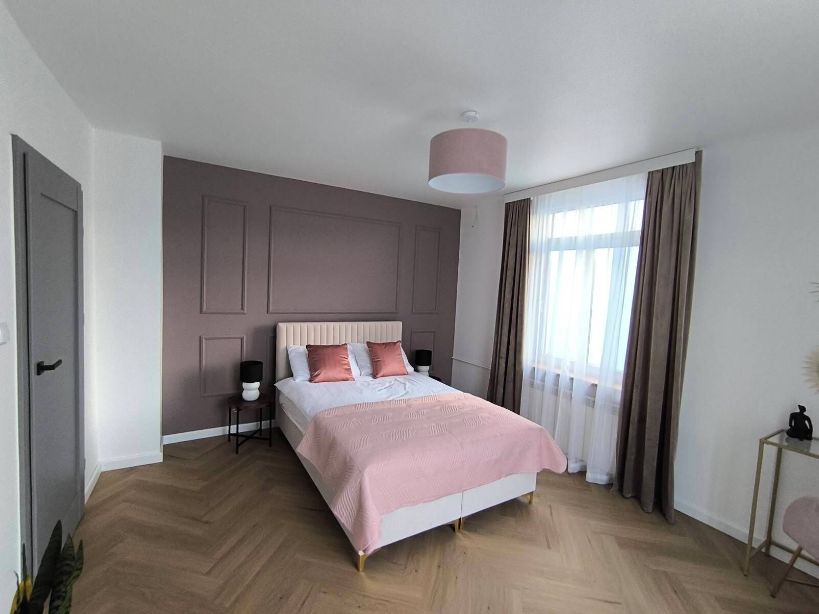 Un dormitorio con cama, mesitas de noche, lámpara, ventana con cortinas y puerta.