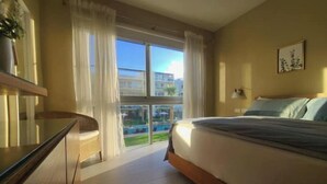 A hotel room with a bed, a desk, a chair, a window with curtains, and a view of buildings and a pool. - vr207h+c. Lovely apartment with pool view.

 (Bayahibe)