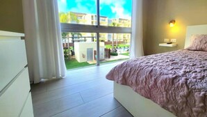 A bedroom with a large window, a bed with a pink blanket, a bedside table with a lamp, and a view of a courtyard with buildings and greenery. - Fantastic view of the pool vr222 h+c
 (Bayahibe)