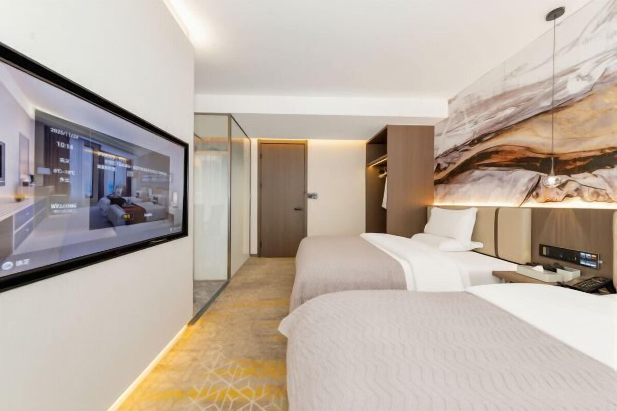 Moderni hotellihuone, jossa on suuri sänky, yöpöytä, seinälle asennettu televisio ja suuri abstrakti taulu sängyn yläpuolella.