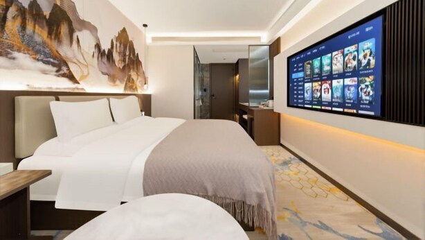 Una habitación de hotel moderna con una cama grande, un televisor montado que muestra una selección de películas y una obra de arte en la pared que representa un paisaje montañoso.