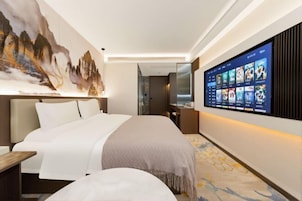 Una habitación de hotel moderna con una cama grande, un televisor montado que muestra una selección de películas y una obra de arte en la pared que representa un paisaje montañoso.
