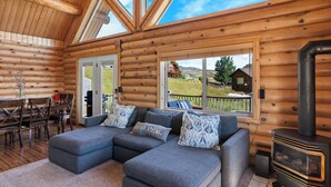 L’intérieur d’une cabane en rondins, avec un canapé d’angle, une cheminée et de grandes fenêtres donnant sur le paysage extérieur.