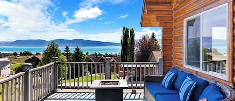 Terraza de madera con sofá de mimbre y un pozo de fuego, con vistas a un lago y montañas.
