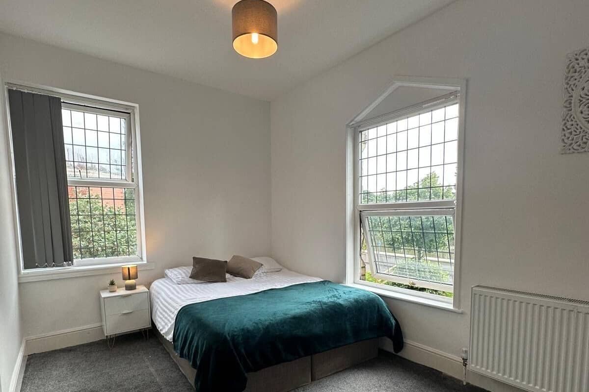 A bedroom with a bed, two windows, a nightstand, a lamp, and a radiator.