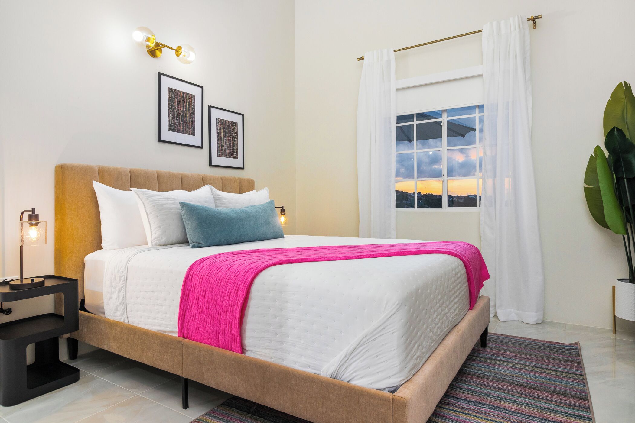 Una cama bien hecha con un edredón rosa, una lámpara de noche y dos cuadros en la pared.