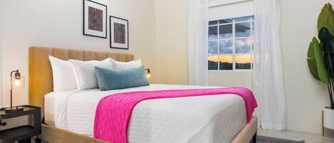 Una cama bien hecha con un edredón rosa, una lámpara de noche y dos cuadros en la pared.