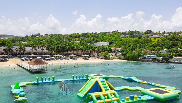 A beach with water play structures, a sandy beach, and a backdrop of buildings and trees. - Sunset at ArecaHomes~ Beach Access~ Pool & Water Park (Discovery Bay)