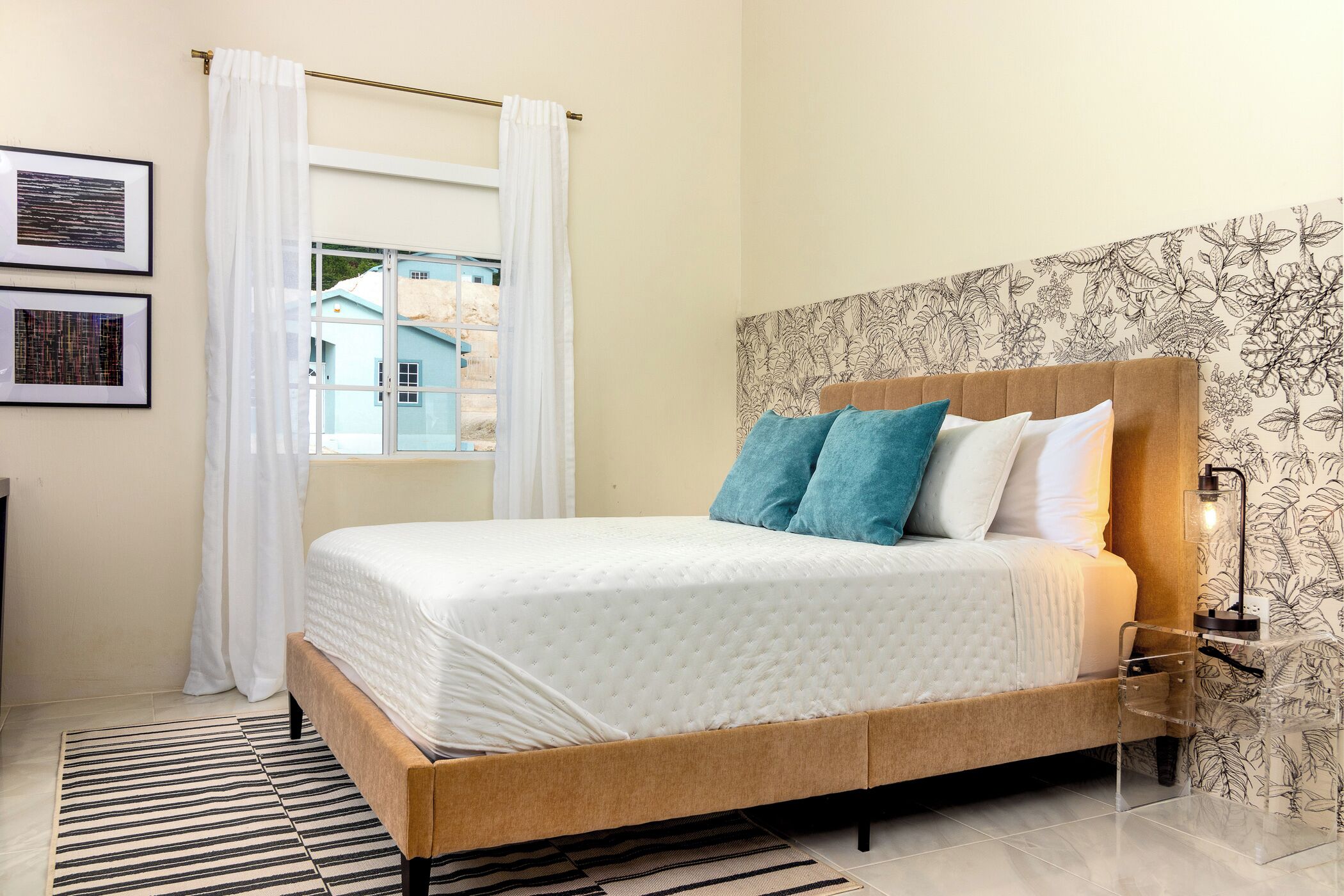 A bedroom with a bed, white curtains, a patterned wall, and a window with a view.